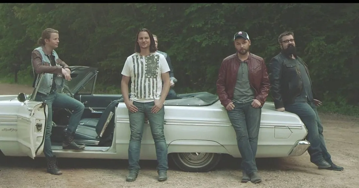 Five individuals stand and sit around a vintage white convertible car in a forested area.