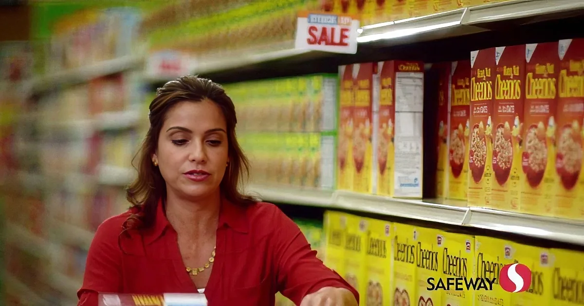 A woman in a red shirt shops for Cheerios at Safeway, surrounded by cereal boxes.
