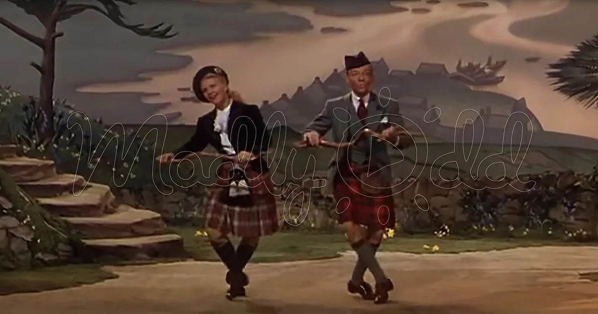A man and woman in kilts dance joyfully on a stage with a scenic backdrop.