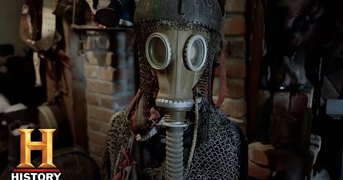 A person wearing a gas mask and patterned clothing stands in a cluttered room with various items.