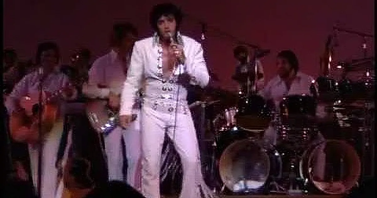 Elvis Presley in white jumpsuit singing on stage with band.