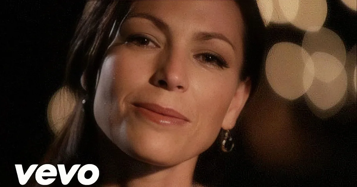 A woman with dark hair and makeup, wearing earrings, smiles slightly against a blurred background of lights.