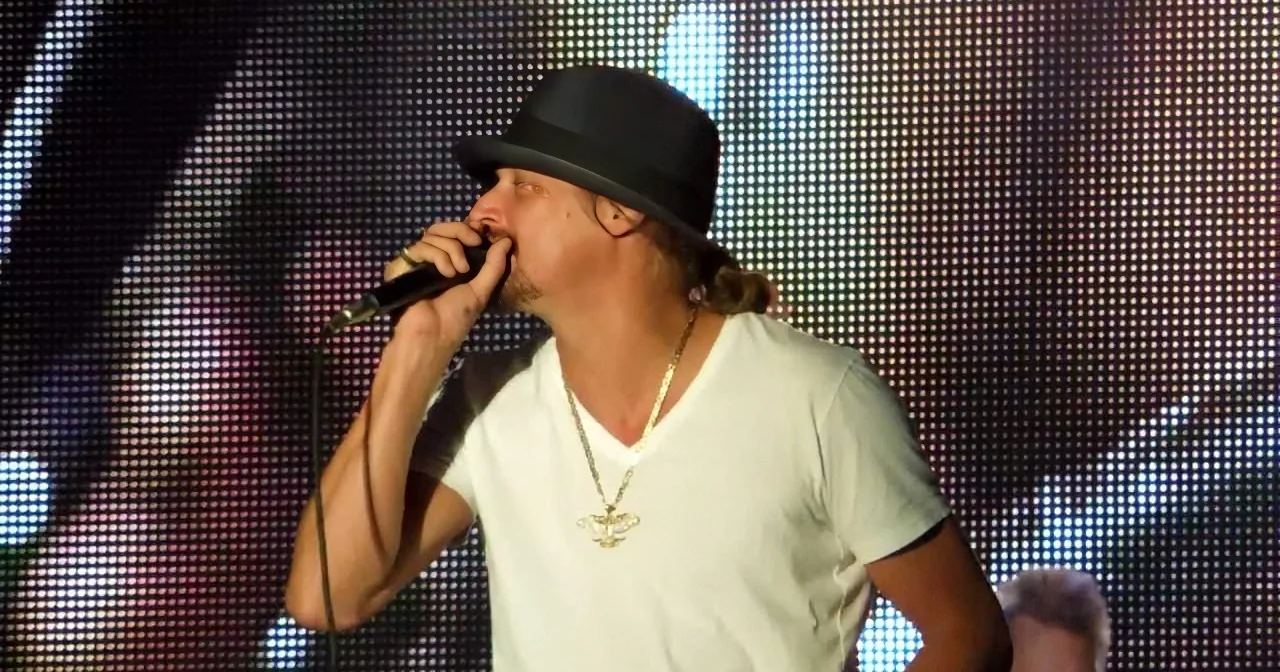 A man in a white shirt and black hat sings into a microphone on stage with a LED screen backdrop.