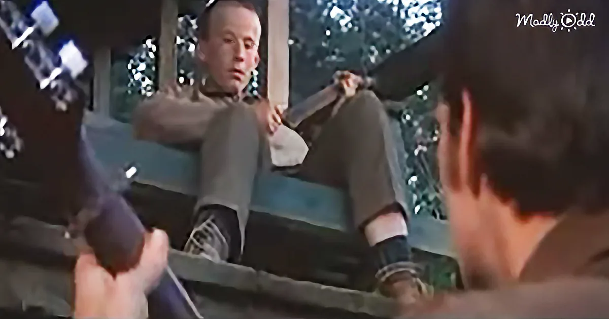 A man sits on a porch swing, holding a gun, while another person approaches.