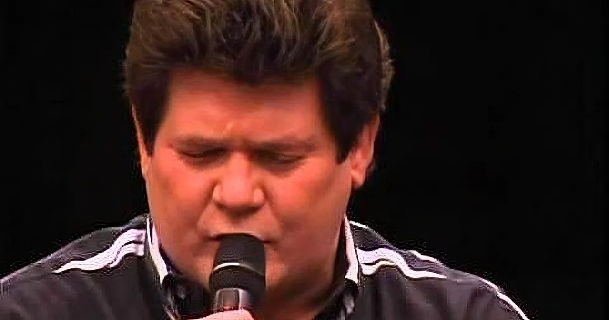 A man with short brown hair, wearing a black and white striped shirt, sings into a microphone against a dark background.