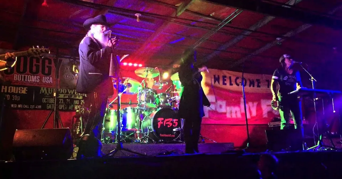 A man with a long white beard plays a harmonica on stage, surrounded by musicians and colorful stage lights.