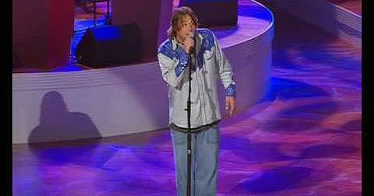 A person in a denim jacket sings into a microphone on a stage with colorful lighting.
