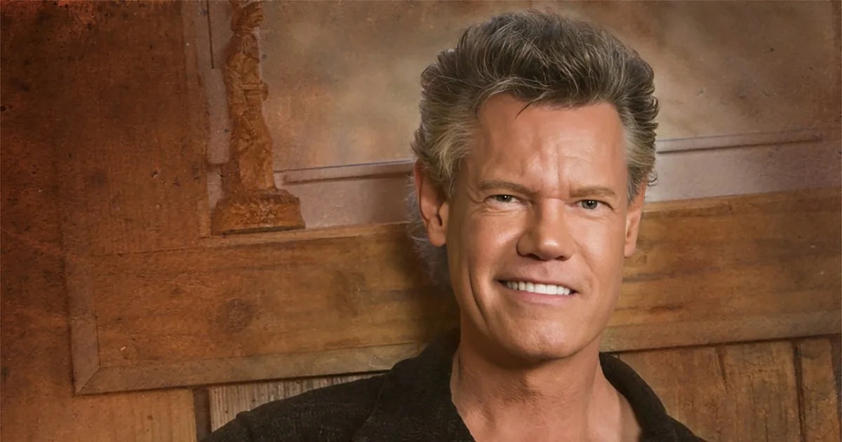 A smiling man with styled hair, wearing a black shirt, stands against a wooden background.