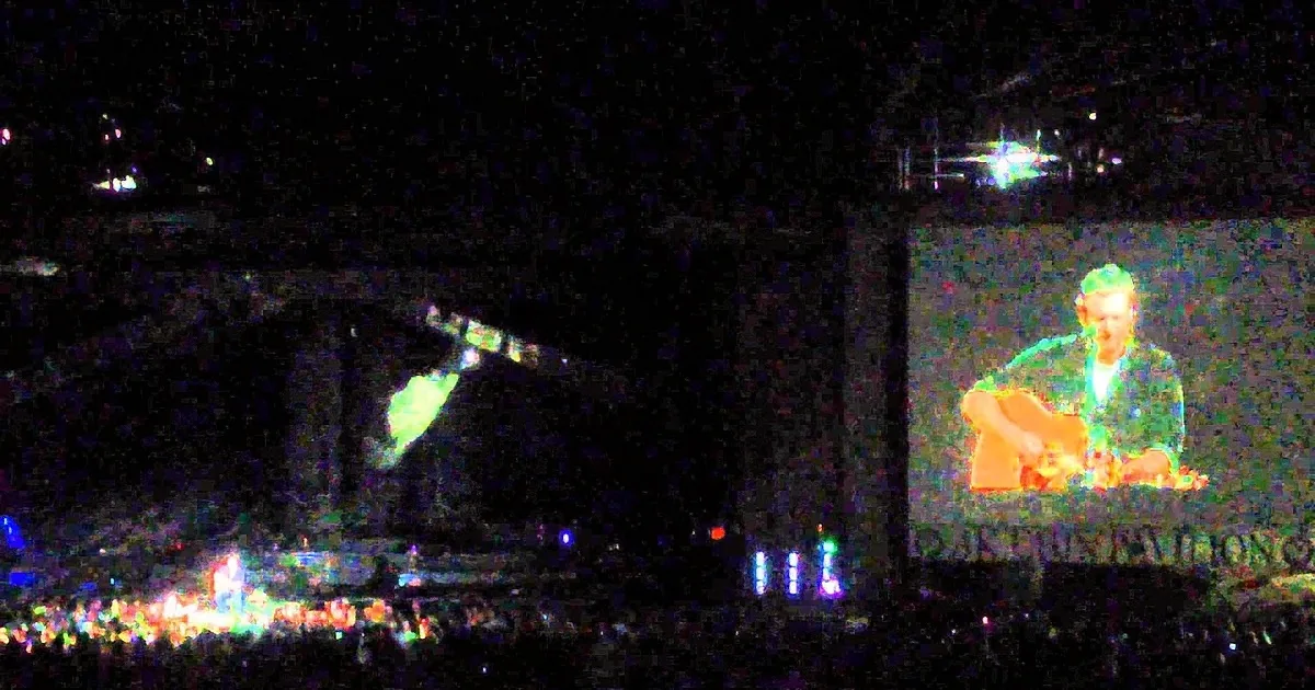 A person plays a keyboard on stage with a large screen showing them, surrounded by a crowd and colorful lights.