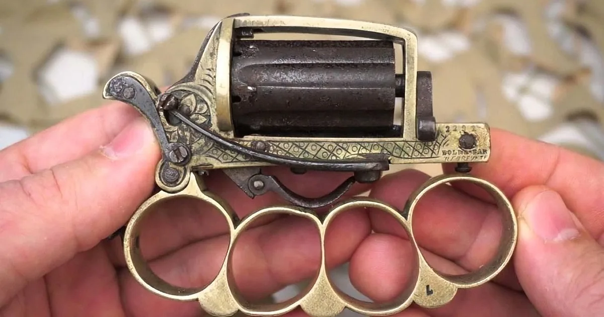 A hand holds a brass knuckle revolver with intricate engravings and a black cylinder.