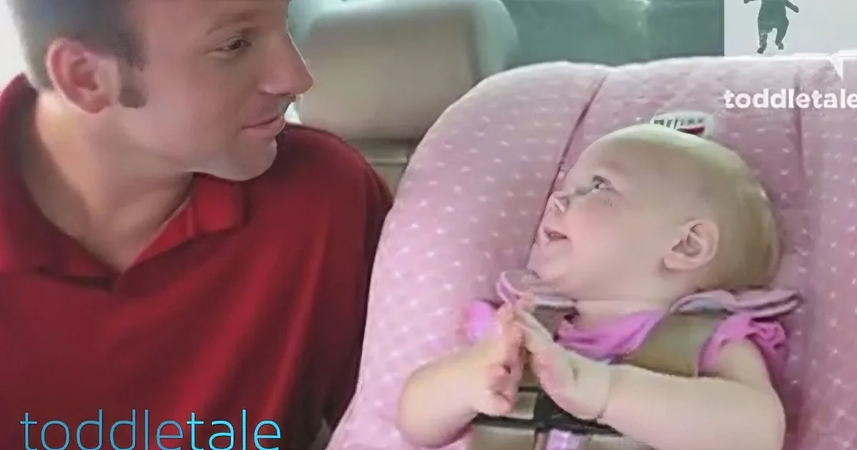A man in a red shirt interacts with a baby in a pink high chair, both looking at each other.