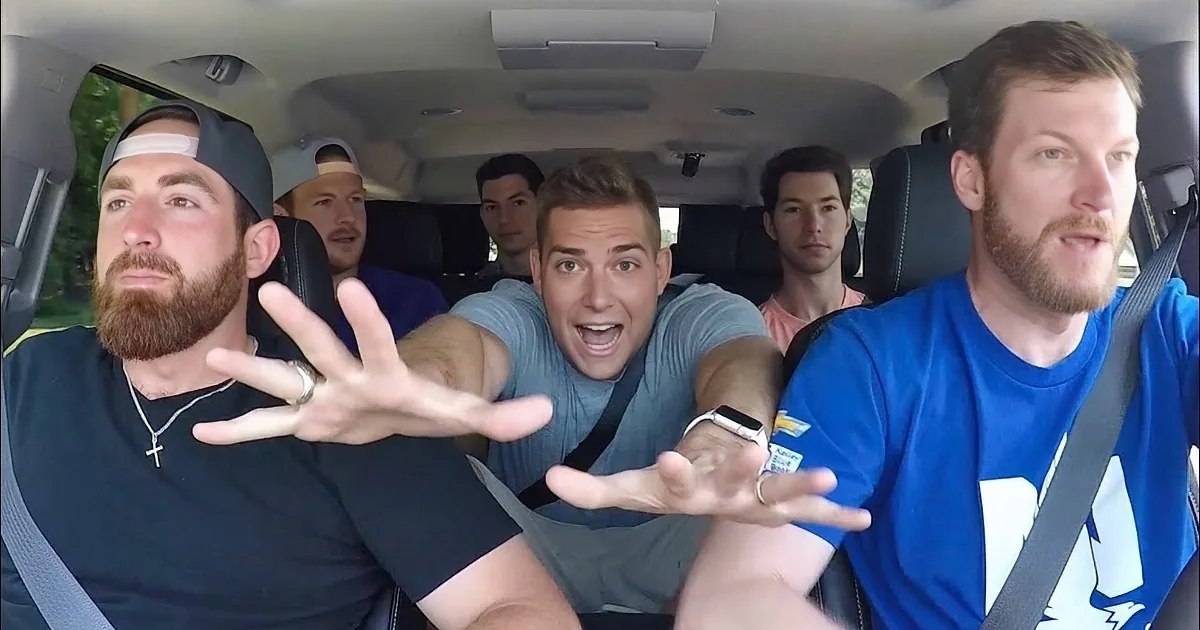 A man in a blue shirt is driving a car, gesturing with his hands while surrounded by other men.