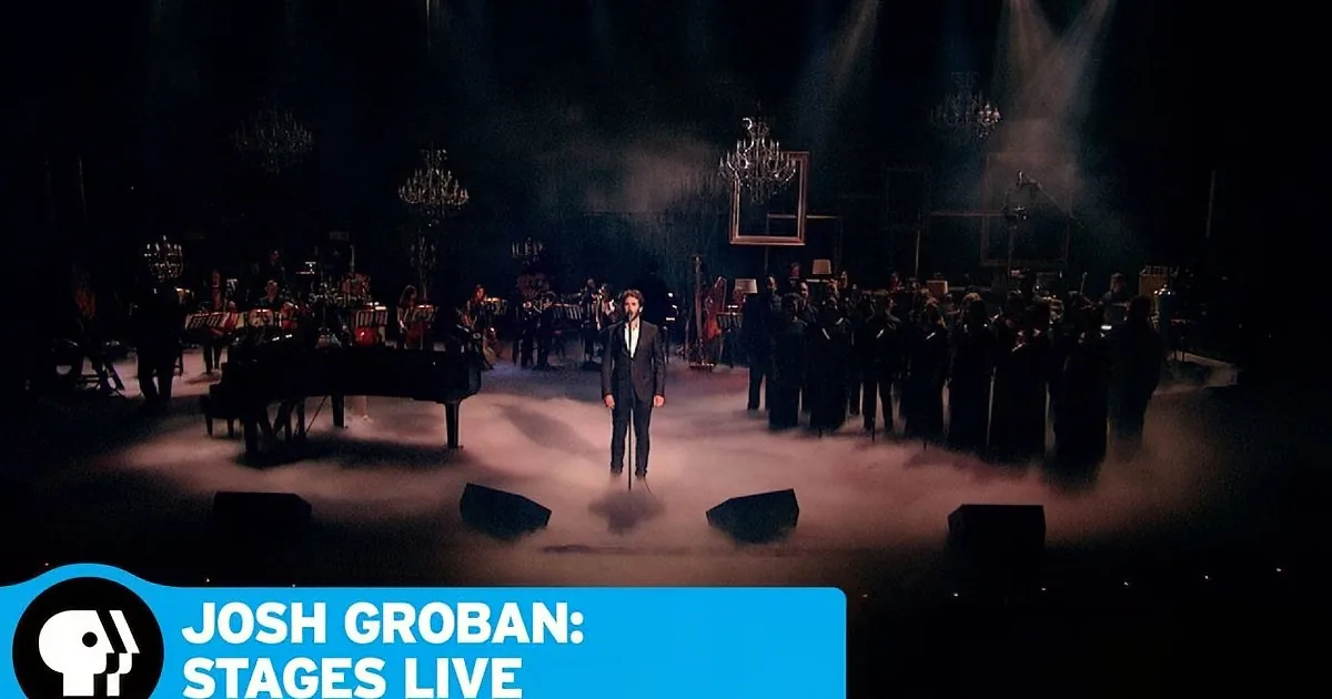 Josh Groban stands center stage with an orchestra and choir behind him, illuminated by spotlights.