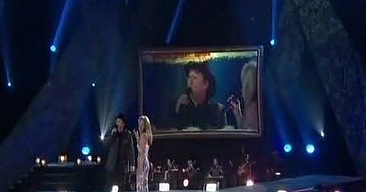 A woman in a white dress sings on stage, with a large screen behind her showing a man singing.