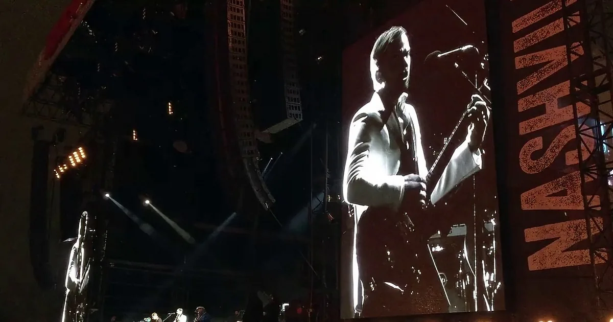 A large screen displays a man playing guitar, with a band performing on stage below.