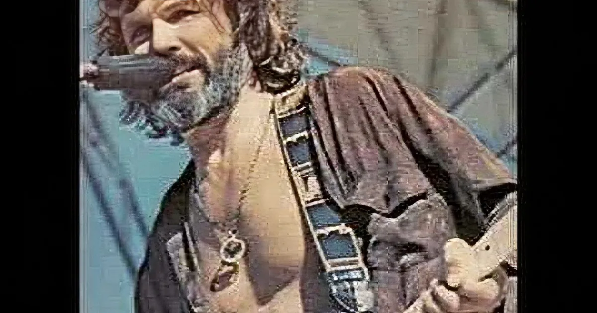 A man with long hair and a beard, wearing a patterned shirt and sunglasses, sings into a microphone.