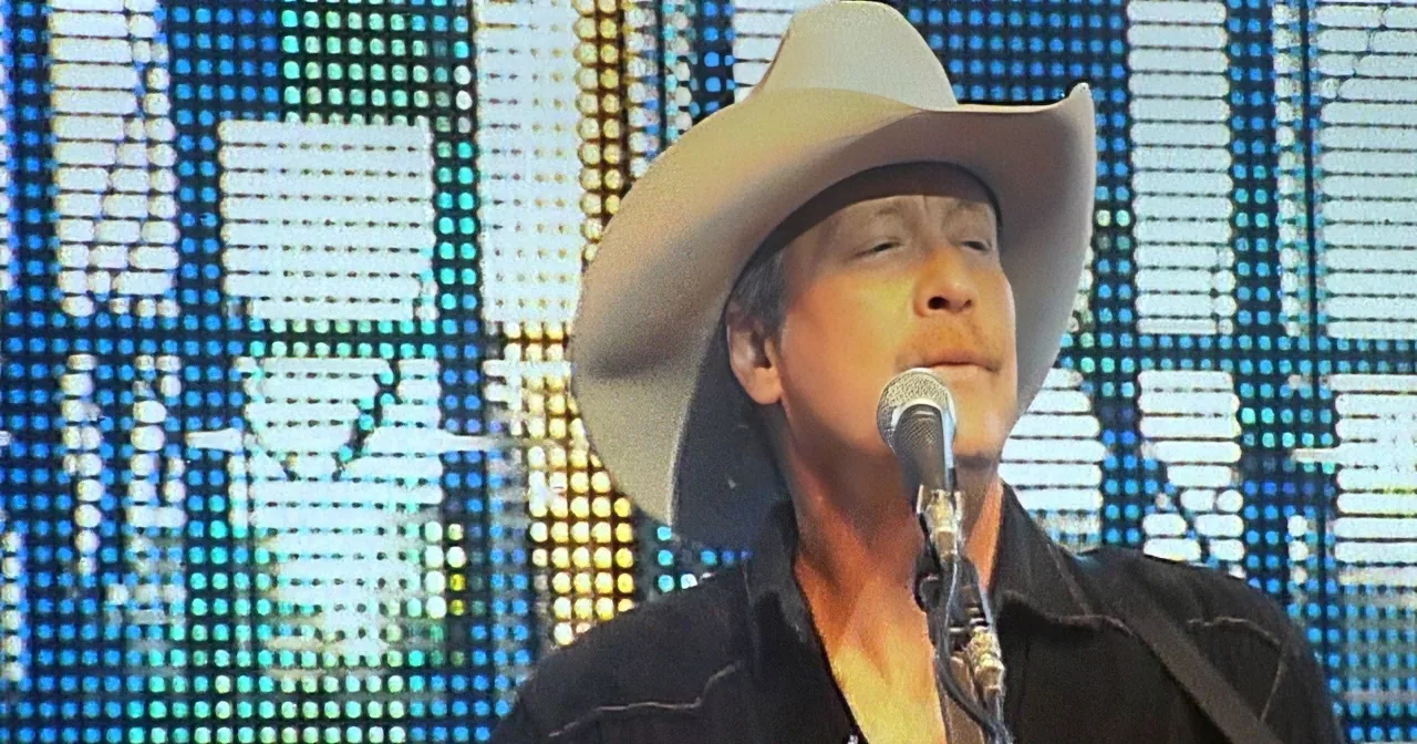 A man in a cowboy hat sings into a microphone on stage with a colorful LED backdrop.