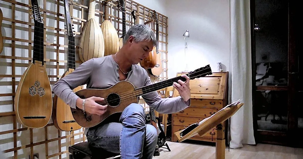A man sits playing a guitar in a room filled with various stringed instruments.