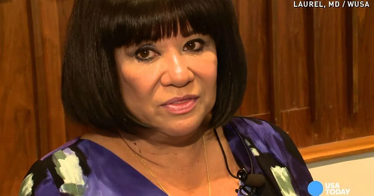 A woman with short dark hair, wearing a purple patterned top and a gold necklace, is speaking in an interview setting.
