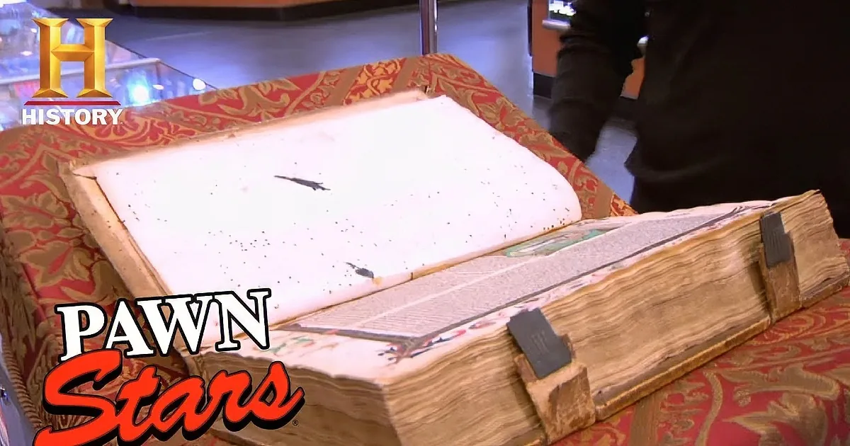 A large, old book with a wooden binding sits on a red patterned tablecloth.