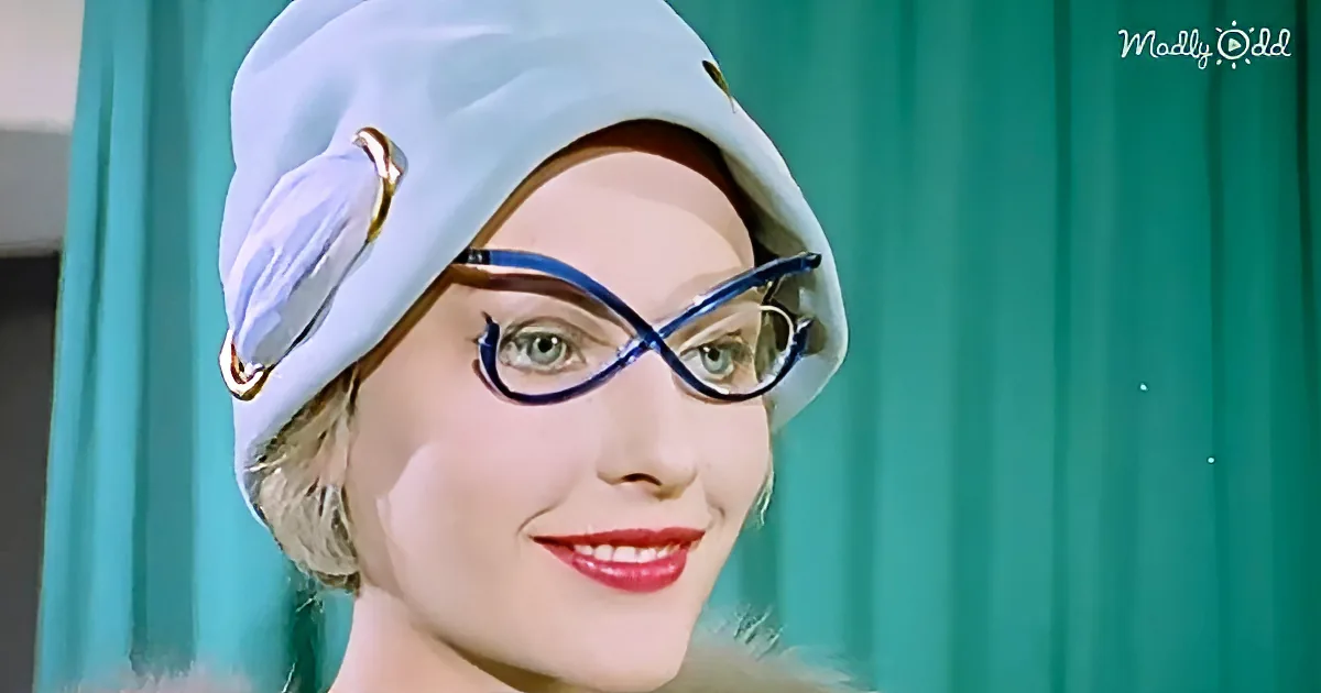 A woman with short blonde hair, wearing a light blue hat and blue-framed glasses, smiles against a teal curtain backdrop.
