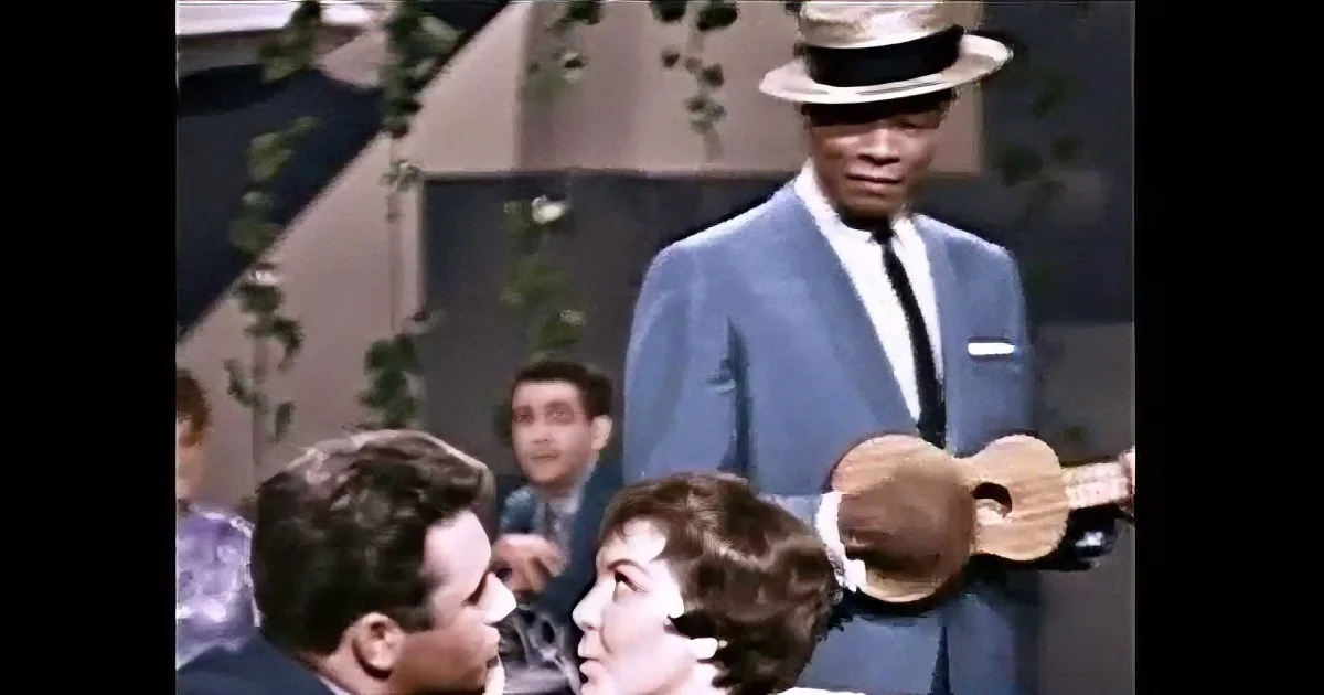 A man in a suit and hat stands holding a ukulele, surrounded by an audience.