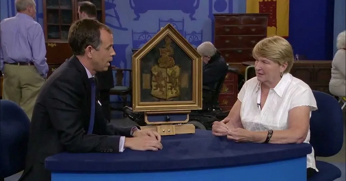 A man and woman sit at a table, discussing an ornate framed object, with a blue backdrop and other people in the background.