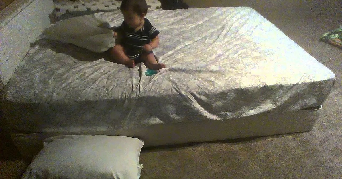 A baby sits on a large, wrapped mattress on the floor.