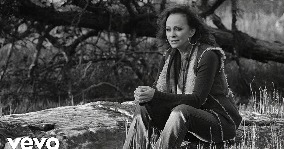 A woman sits on a log in a forest, wearing a fur-trimmed vest and jeans, surrounded by trees.
