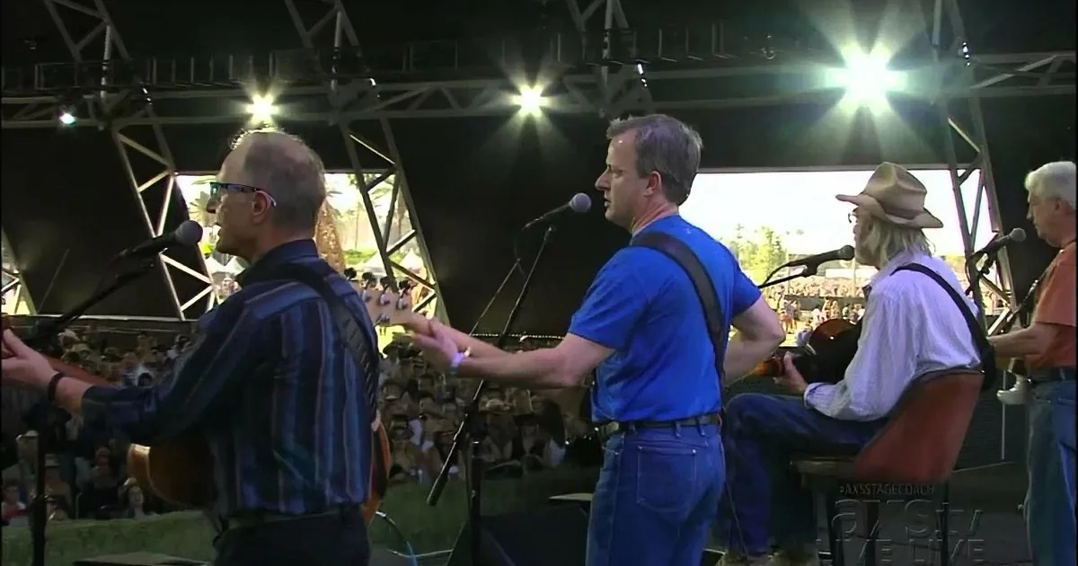 A band performs on stage with microphones, guitars, and a crowd in the background.