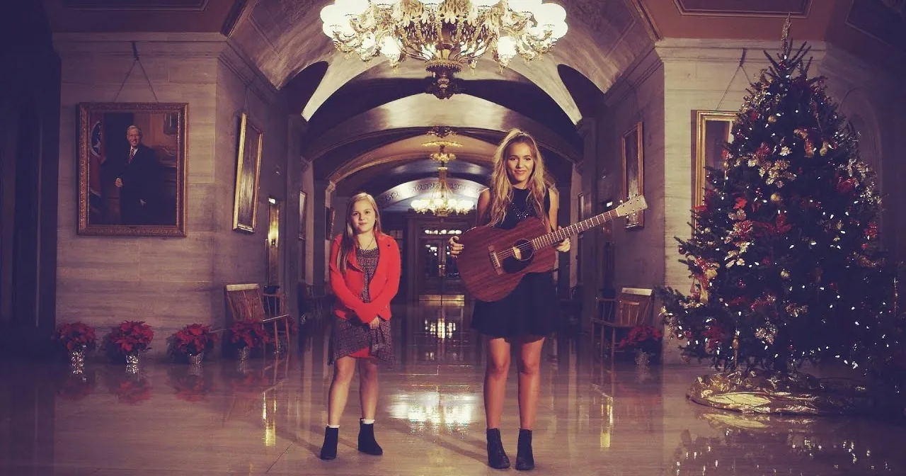 A woman holds a guitar in a grand hall with a Christmas tree and portraits, reflecting festive and formal ambiance.