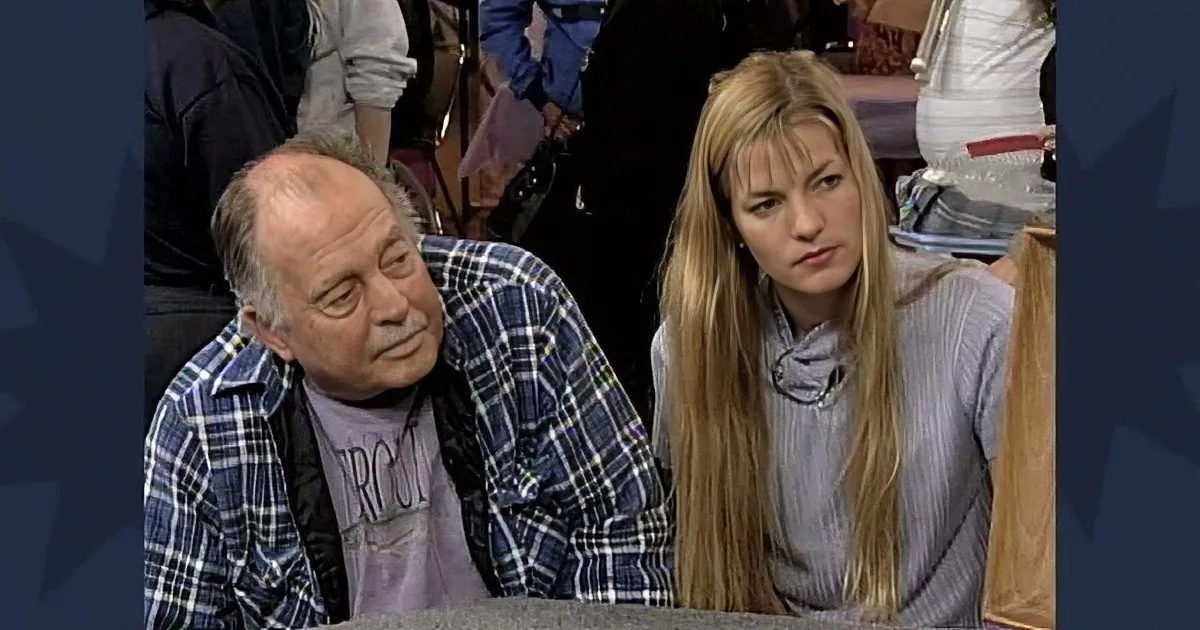 A man in a plaid shirt leans on a table, while a woman with long blonde hair sits beside him, both appearing to be in a...