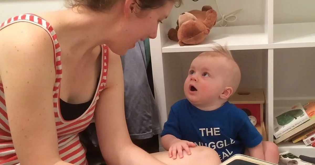 A woman in a red and white striped tank top is reading to a baby in a blue shirt, who looks surprised.