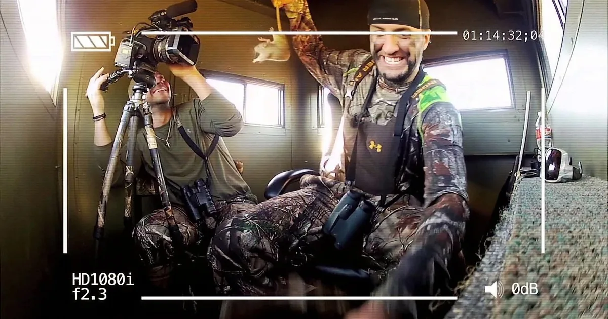 A man in camouflage gear is holding a small animal, smiling, while another man films with a camera.