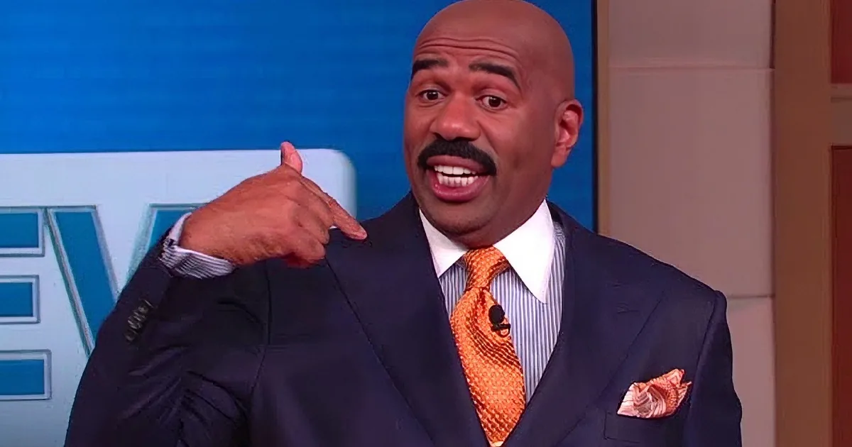 A man in a suit and orange tie is gesturing with his hand, smiling.