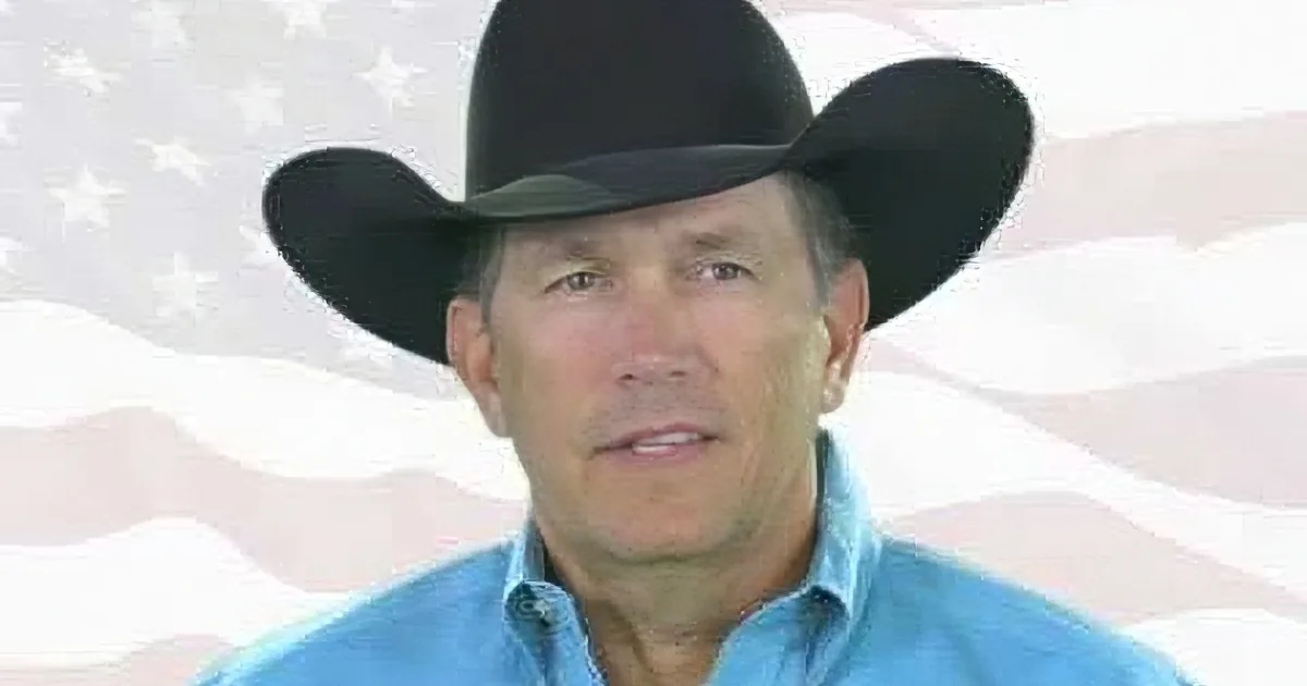 A man in a cowboy hat and blue shirt stands in front of an American flag.