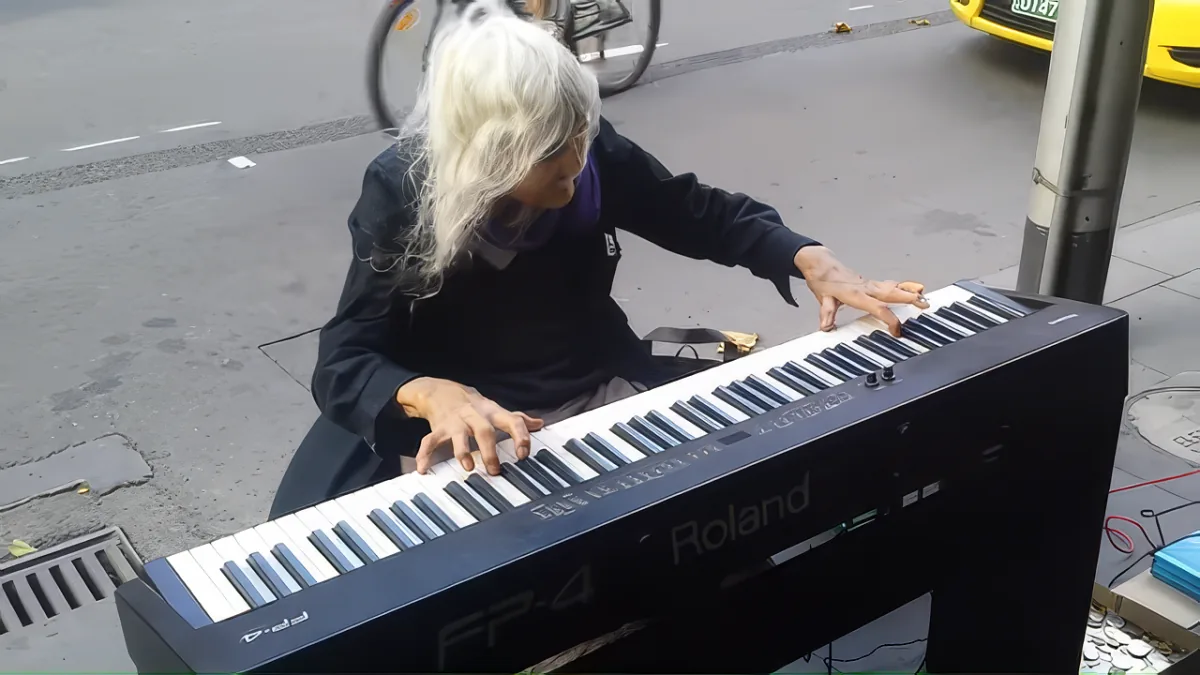 An elderly person plays a keyboard on a city street, with a bicycle and car in the background.