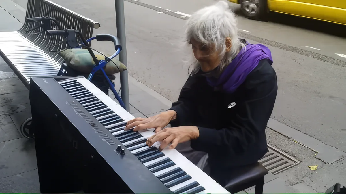 An elderly woman plays a keyboard on a city street, surrounded by urban elements.