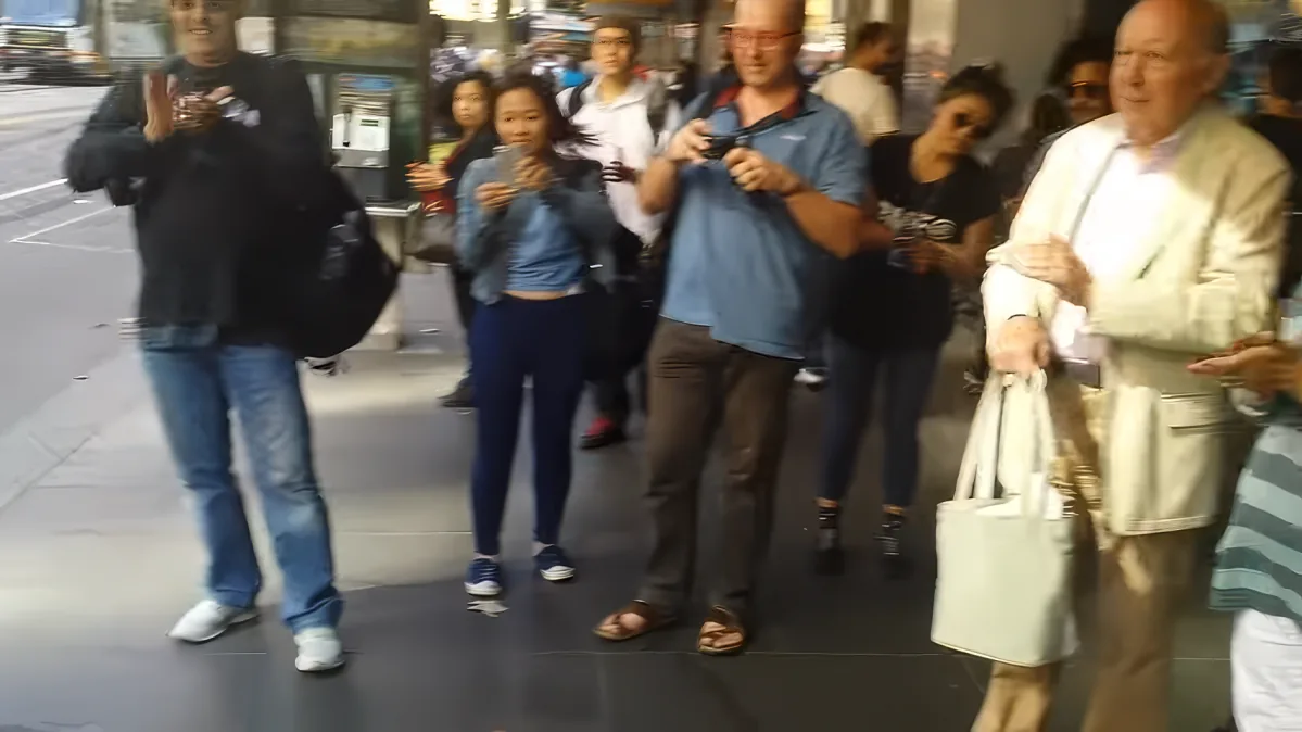 A man in a blue shirt and sandals holds a camera, walking with a group of people on a busy street.