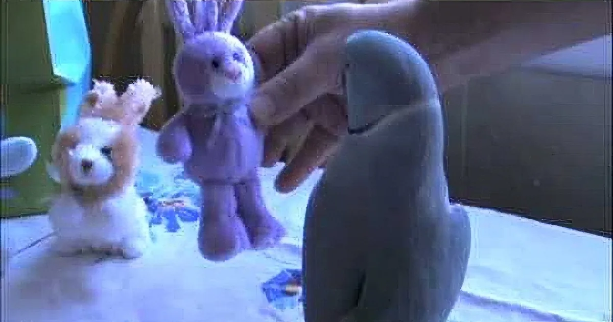A hand holds a purple stuffed bunny, surrounded by other plush toys on a table.