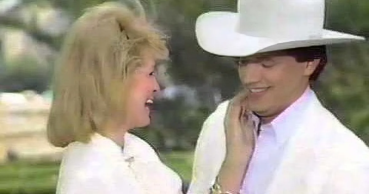 A man in a white cowboy hat and a woman in a white blouse are embracing, with the man's hand on the woman's face.