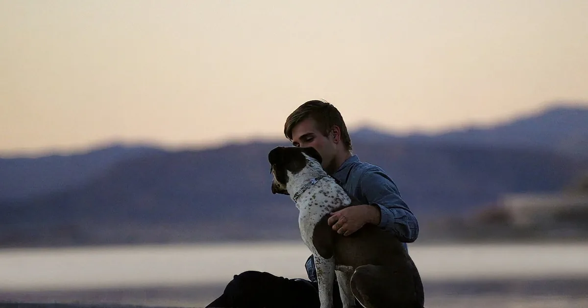 A man hugs a dog at dusk, with mountains and water in the background.
