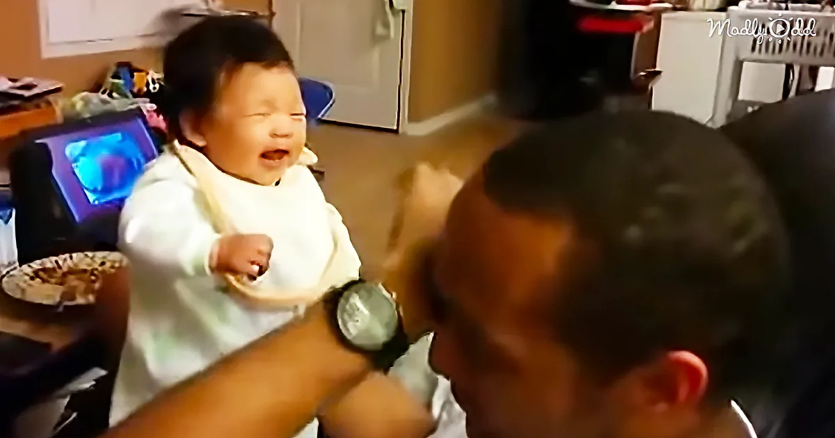 A baby in a bib is laughing while a person in a watch holds a spoon near them.