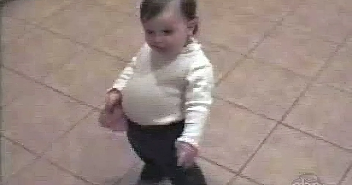 A baby in a white sweater and dark pants stands on a tiled floor, looking down.