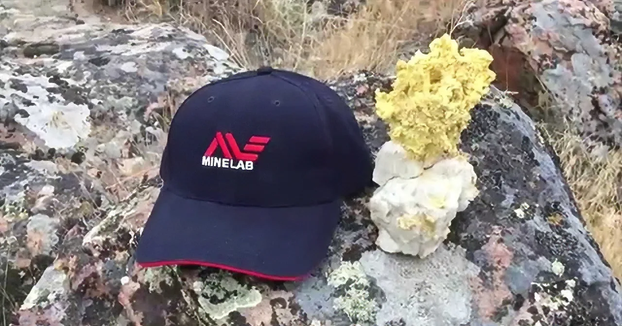 A black Minelab-branded cap lies on a rocky surface next to a yellowish mineral formation.