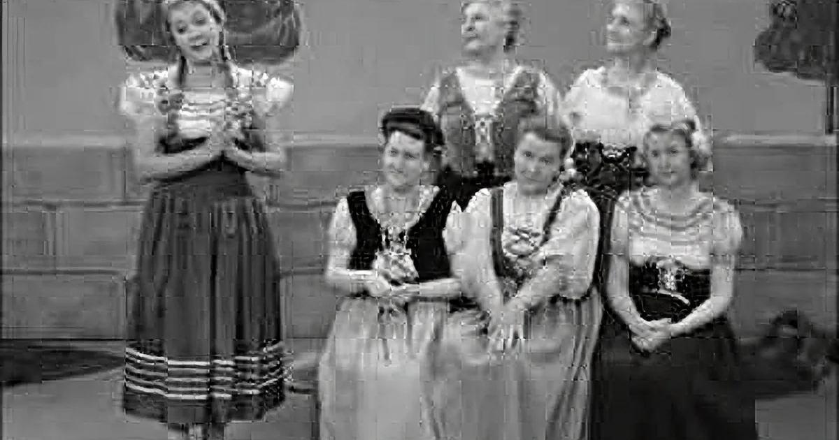 Women in traditional attire stand in a line, some holding hands, against a backdrop resembling a stage or set.