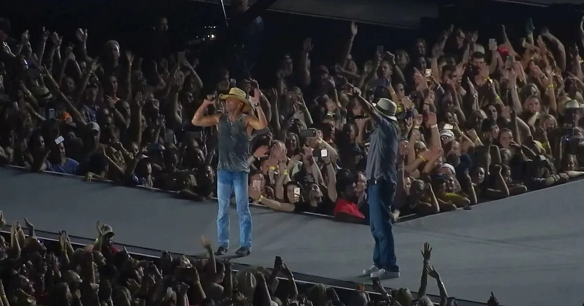 Two men on stage, one in cowboy hat, crowd cheering, hands raised, bright lights.
