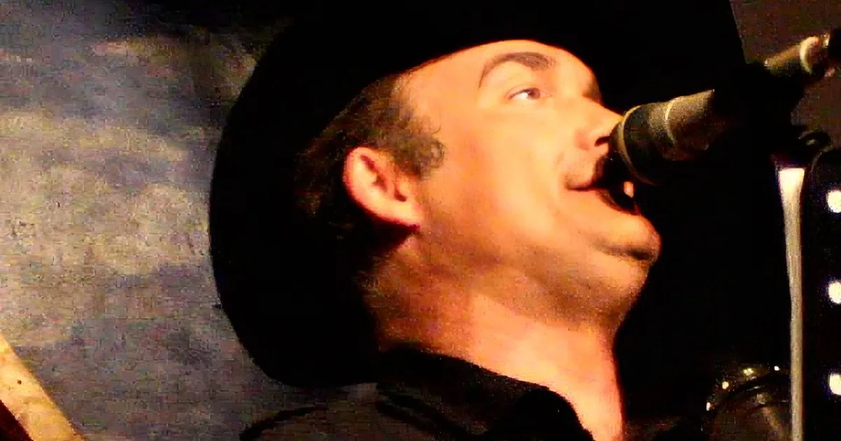 A man in a black hat sings into a microphone, wearing a dark shirt, with a blurred background.
