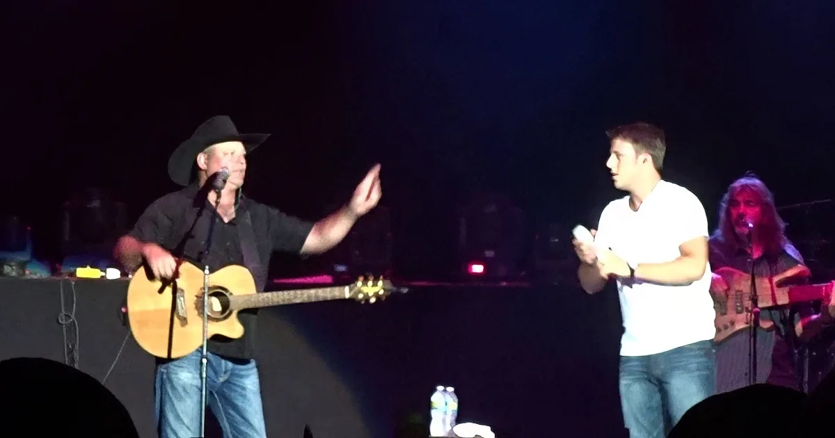 A man in a cowboy hat gestures while another man holds a microphone on stage.