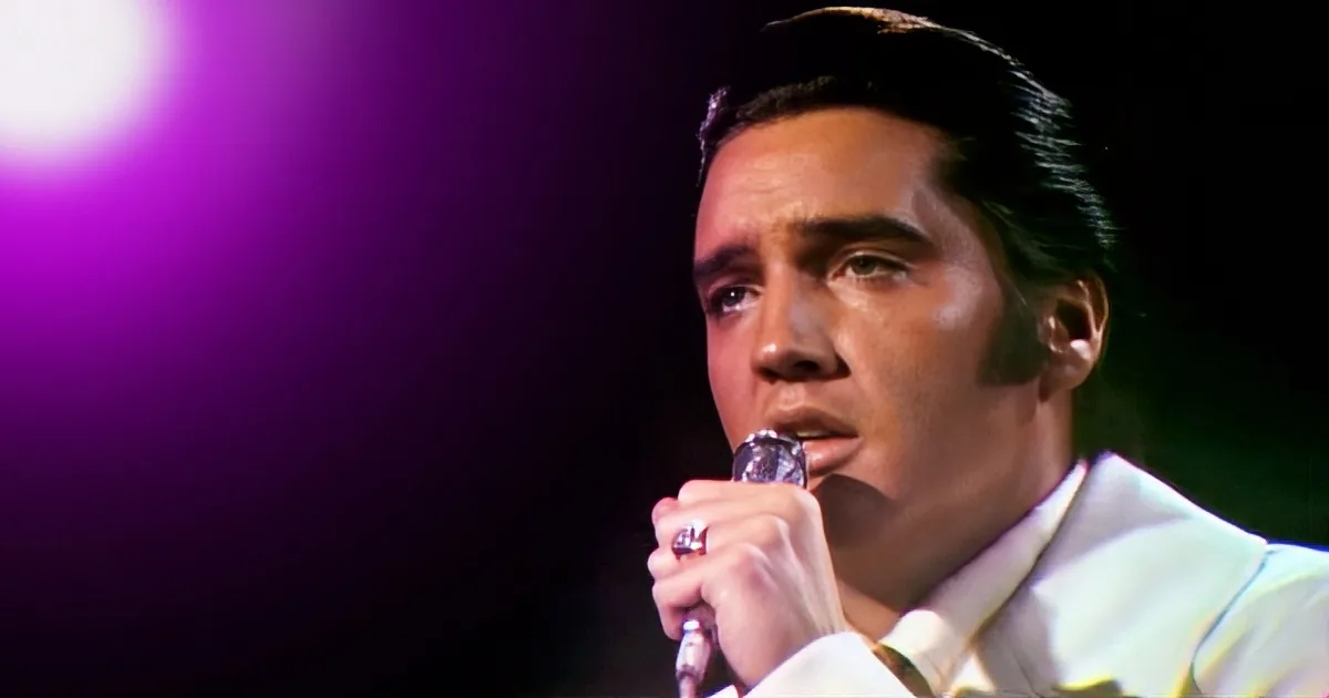 A man in a white suit holds a microphone, singing with a purple light behind him.